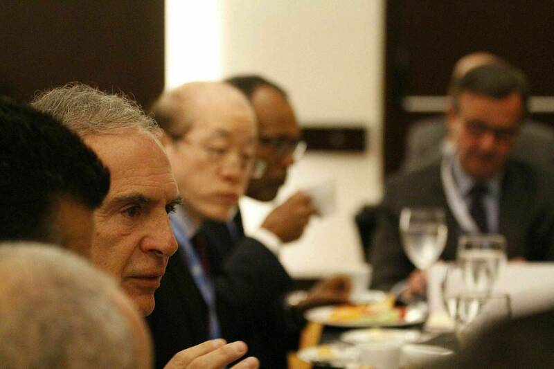 A man with short gray hair gestures while speaking at a table during a meeting. Other attendees in business attire sit around the table, some eating and drinking.