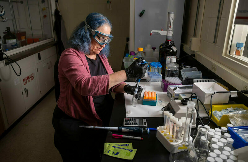 A researcher with blue-tinged hair and safety goggles uses a pipette, wearing black gloves while working at a lab bench amongst various scientific equipment.