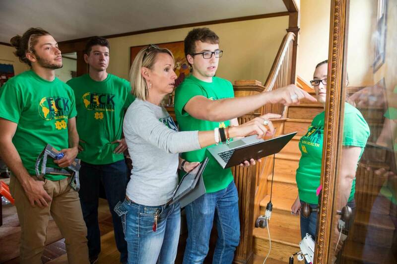 A woman and four students wearing Eck Institute for 全球 Health t-shirts review information on a laptop and tablet next to a wooden staircase.