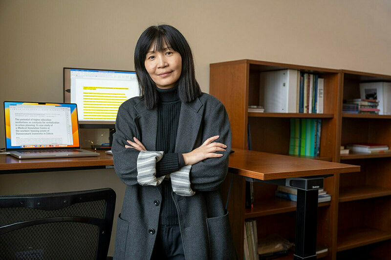 An Asian woman with shoulder-length dark hair and a gray coat stands facing the camera with arms crossed in front of a sit-stand desk. Two computer screens and a bookshelf are visible in the background.