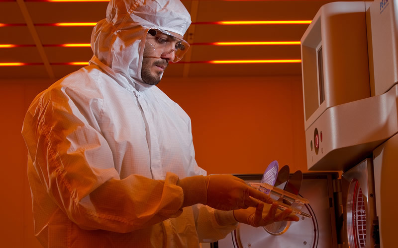 A researcher in a full-body protection suit holds a petri dish by a machine.