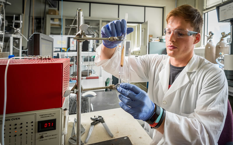 A researcher in a white lab coat and goggles holds a vial near a machine.