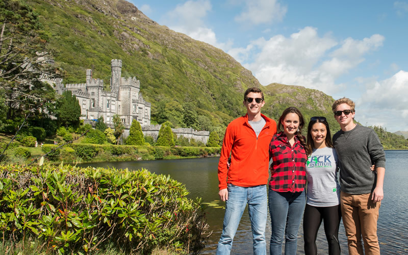 Four students pose in front of Kylemoore Abbey, with lush green bushes and a lake.