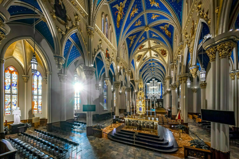 Interior view of the Basilica of the Sacred Heart at the 圣母大学. Sunlight streams through stained-glass windows. The ornate gold altar and pipe organ are visible. Rows of chairs are set up on the polished floor. The arched ceiling is painted blue with gold stars and angels.