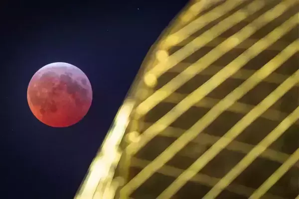 A reddish-orange moon during a lunar eclipse against a dark sky, partially obscured by the blurred Golden Dome.