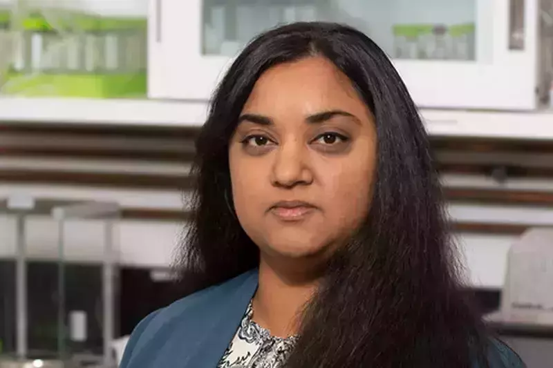 A person with long dark hair and a teal jacket looks at the camera in front of lab cabinets.