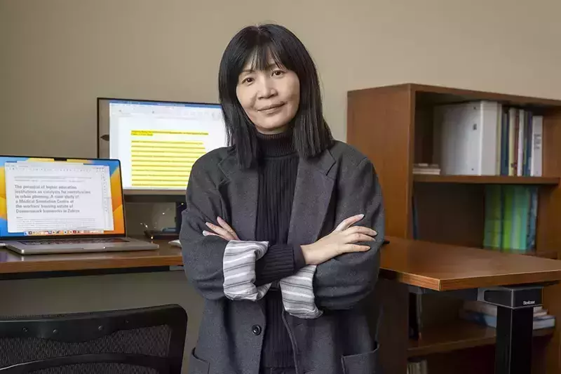 A person with shoulder-length dark hair, wearing a dark gray blazer over a black turtleneck, stands with arms crossed in front of a sit-stand desk. A laptop and monitor displaying academic text are visible on the desk, and a bookshelf is in the background.