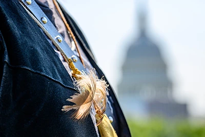 Close-up of a person wearing a dark jacket adorned with light brown feathers and a tan leather strap, with the out-of-focus Capital Building in Washington D.C. in the background.
