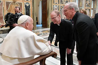 TPope Francis, while seated in an ornate chair, shakes hands with Fr. Bob Dowd. Fr. John Jenkins stands beside them, and a cameraman films the interaction in a grand, marble-floored hall.