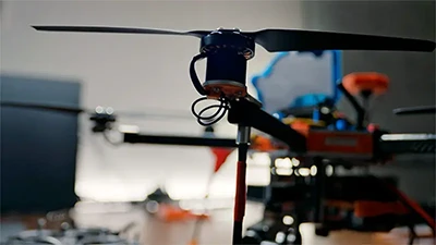 Close-up of a drone's rotor and arm. The black rotor blades are slightly blurred, suggesting motion. Black and orange components of the drone are visible in the background.