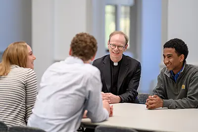 Father Robert A. Dowd, C.S.C., speaking with a group of three students seated at a table.