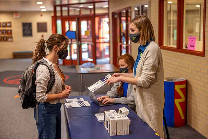A girl wearing a backpack and mask speaks to woman, also wearing a mask and pointing to a sheet of paper, about wifi hotspots.