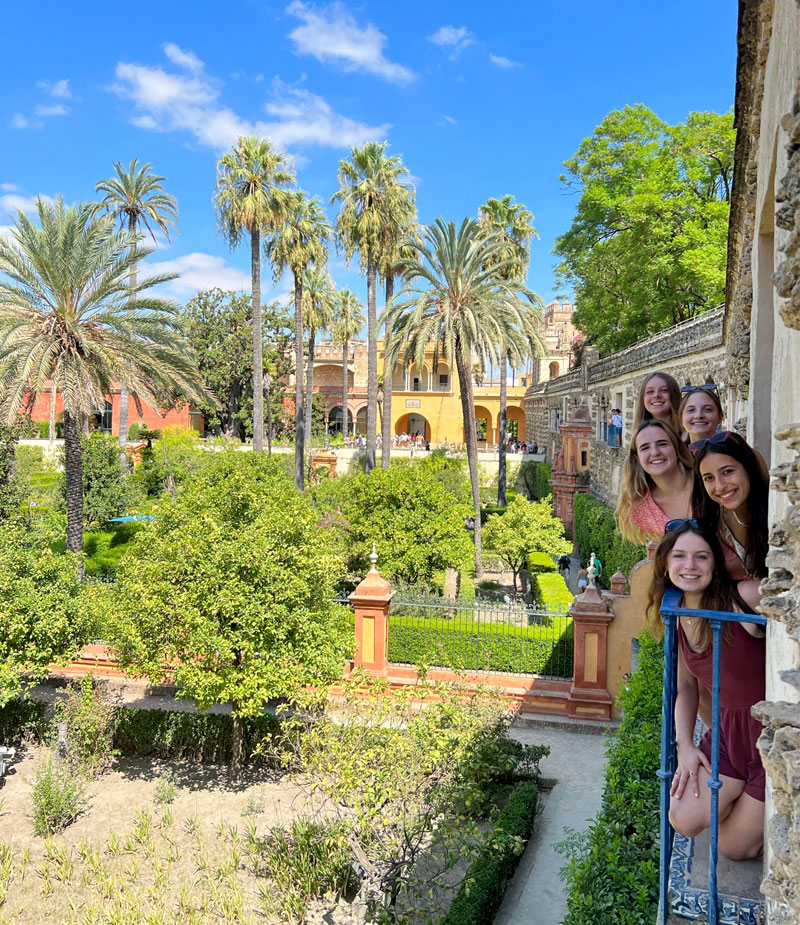 Five female Notre Dame students smile from a balcony window overlooking palm trees and greenery in Alcázar in Sevilla.