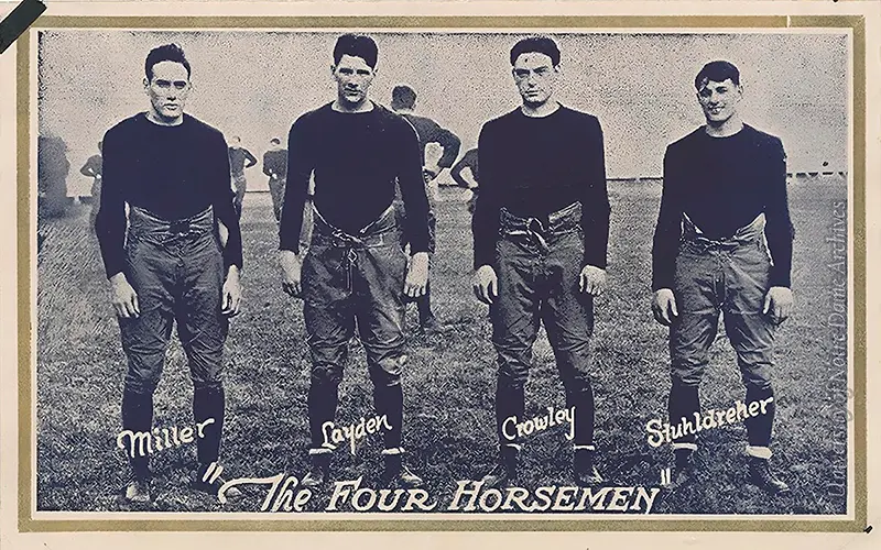 A sepia-toned photo of Notre Dame's 'Four Horsemen' football players. From left to right, they are identified as: Miller, Layden, Crowley, and Stuhldreher. They stand on a field in uniform, other players visible in the background. The words, 'The Four Horsemen' appear below the players.