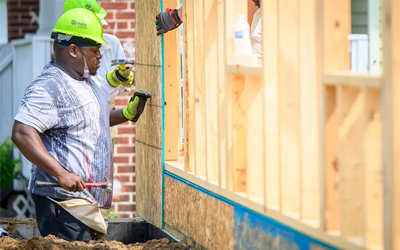 A construction worker wearing a lime green Habitat for Humanity hardhat uses a hammer to build the frame of a house. He's wearing work gloves and holding the hammer in his left hand.