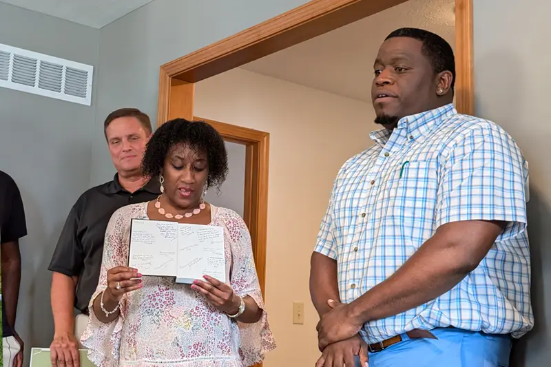 A woman reads aloud from a notebook filled with handwritten messages. A man stands listening behind her to her left, and another man stands to her right with his hands clasped. They appear to be inside a house or office.