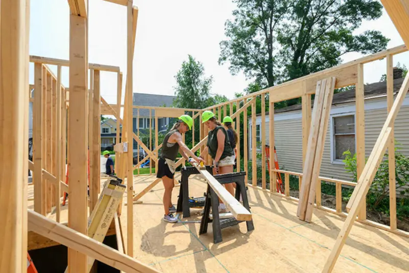 Three women wearing neon green hard hats stand at a saw in the middle of a house frame, 